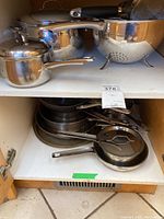 Cookware set stored in cupboard, showing a small saucepan with lid, various pots and pans stacked on shelves.