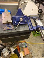 Wide view of the metal anvil and two blue metal bench vises on a workbench in a garage environment.