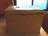 Square brown saddle faux leather tufted storage ottoman bench with hinged lid, shown closed, placed near window on wood floor.