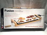 Front view of the Swissmar Fusion box showing ceramic top table grill with food on the platter and basic product features.