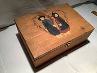 Top and side angled view showing wooden box with hand painted Pilgrim boy and girl dolls inside a heart shape on lid.