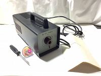 Main view of the vintage Russian microscope light source device showing the black metal casing, front panel with circular fiber optic connection port, attached power cable, screwdriver tool, colored glass bulb, and instruction pamphlet.