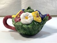 Side view of small vintage ceramic teapot showing colorful flowers, green leaves, red handle, and spout.
