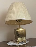 Pair of brass floral etched lamps with cream pleated shades and clear plastic covers, sitting on a lace doily.