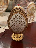 Front view of Faberge egg replica showing intricate gold-toned and silver-toned exterior with beaded and jewel accents, sitting on ornate gold-colored base.