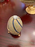 Photo shows gold-colored Faberge egg replica with blue gems arranged in spiral pattern and a large clear gem on top, standing on gold-tone ornate three-legged base.