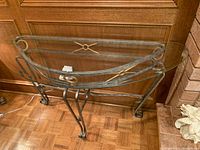 Glass and metal half-moon-shaped console table photographed against a wooden paneled wall and hardwood floor, showing metal legs with scrollwork and gold-tone decorative hardware accents.