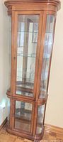 Whole view of curved oak curio display cabinet with two glass doors and glass shelves, upper cabinet includes light.