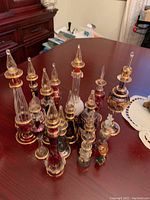 Group of 11 decorative perfume bottles of various colors, sizes and intricate gold detailing with pointed stoppers on wood table