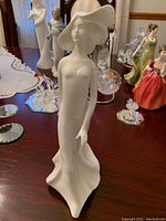 Front view of the white porcelain figurine showing a woman in a flowing dress and wide-brimmed hat on a wooden table with other figurines in the background.