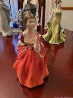 Front view of the Royal Doulton Flower of Love figurine showing a woman in a red dress holding a pink flower, positioned on a wooden surface with other figurines in the background.
