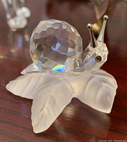 Close-up photo of Swarovski crystal snail figurine on a frosted crystal vine leaf base, showing faceted crystal shell and detailed texture of leaf.