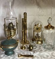 Photo showing a collection of brass and brass-colored decorative items including oil lamp, mantle clock with cracked dome, lidded jar, ceramic urn, small brass teapot and cigarette lighter.