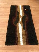 Full view of hand hooked rectangular wall hanging or mat with central cream/gold/olive design on dark brown background on wood floor.