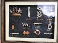 Close look at first shadow box displaying multiple maritime knots with labeled plaques, small wooden ship model and decorative spool and life ring.