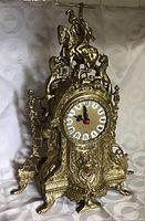 Front view of ornate solid brass clock with detailed embossed decorations and horseman figure on top. Clock face has Roman numerals and black hands with second hand red.