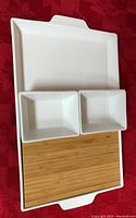 White porcelain tray holding a removable bamboo cutting board and two small white rectangular bowls, shown on a red cloth background.