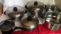 Photo showing eight pots with lids, includes Cook o Matic pots with black handles and knobs, a smaller saucepan with black handle, a large pot with glass lid, on red tablecloth and white background.
