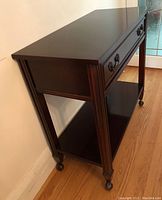 Angle view of dark wood rolling server showing drawer with metal handles, bottom shelf, caster wheels, and vertical panel detailing on legs.