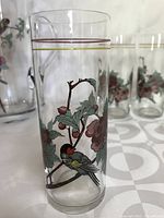 Close-up of a tall glass showing bird on a branch with leaves and pink flowers, with gold trim near the rim.