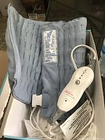Light blue fabric heating pad laid out in box showing attached cord and controller with Sunbeam branding.
