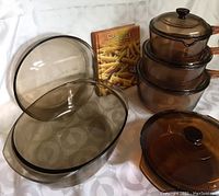 Full view of all items including three stacked Vision Corning glass pots with lids, oval roaster, round casserole dish, and pasta cookbook