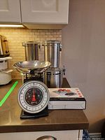 Photo of mechanical Starfrit kitchen scale with large stainless steel bowl and four chrome canisters in background with original Imperial Houseware kitchen scale box sitting to the side on kitchen counter.