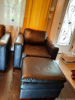 Full view of the dark brown/black leather chair with its matching faux leather ottoman placed in front on wooden floor with yellow curtains in the background.