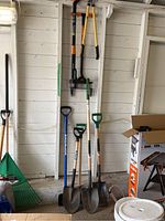 View of all 8 garden tools including 3 shovels, an ice chipper, edger, weeder, pruners, and aerator hanging or leaning against white wooden wall