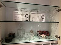Shelf with two boxes of IKEA SVALKA wine glasses, multiple wine glasses displayed, clear decanter, black coasters, whiskey stones, and bottle openers arranged on glass shelf.