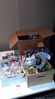 Overall view of the assorted hardware lot showing multiple boxes and bags with various hardware items including boxed wiring accessories, plastic drop sheet packaging, and coiled plumbing hose.