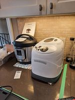 Instant Pot electric pressure cooker and Black & Decker bread maker placed side by side on kitchen countertop with Instant Pot manual behind them.