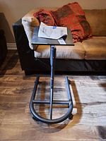 Front view of the metal laptop butler showing the flat rectangular platform and C-shaped base with caster wheels, placed on a wooden floor in front of a sofa.