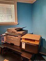Overview shot showing Brother copier, Canon Pixma printer, toner cartridge box, and UniFi access point box on black desk against blue wall.