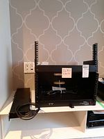 Front view of the black metal rack mount PDU with power cord, mounted on small shelf against wall patterned in wallpaper.