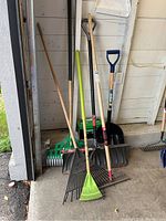 Overview photo showing all six tools including three leaf rakes, one metal rake, and two snow shovels leaning against wall.