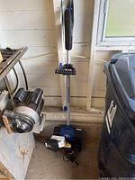 SnowJoe cordless snow shovel standing upright against a wall with battery and charger placed on the floor in front of it.