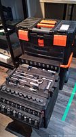 Closed black and orange plastic portable tool box with multiple trays extended showing sockets, ratchets, extensions, and adapters inside
