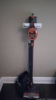 Full upright vacuum standing against the wall with storage bag on the floor next to it.