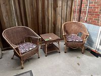 Photo showing two matching vintage wicker chairs with floral seat cushions and a wicker table in between, set against a wooden fence.