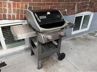 Front and left side view of assembled Weber Spirit II natural gas grill with two stainless steel side shelves, black lid with integrated thermometer, two control knobs, wheels, stands on concrete