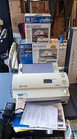 Photo showing Brother Cool Laminator machine with refill packaging boxes stacked behind it, papers on table surface.