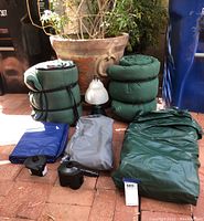 Overview of all camping items including rolled sleeping bags, air mattresses, battery-operated lantern, and air pumps arranged on brick floor.