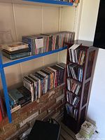 Wooden CD tower filled with CDs and DVDs next to metal shelves with more CDs and DVDs.