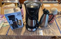Full view photo of all items on a table, showing boxed Primula cold brew coffee maker, DeLonghi coffee maker, and two coffee urns side-by-side.