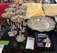 Six silver-plated stemmed glasses, Gorham silver-plated bowl, and Twinkle silver polish kit displayed on black table, yellow cloth in background.