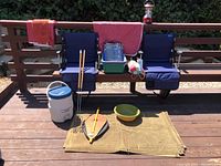 Camping gear lot on wooden bench and deck including two navy blue folding camping chairs with footrests, indoor/outdoor insulated jug, handmade bellows, grilling sticks, green basin, towels, and hammock in bag.