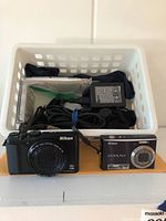 Two Nikon Coolpix cameras in front of a white basket containing various camera accessories including a charger and cables.
