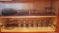 Wooden cabinet shelves containing clear glass plates, bowls, teacups, creamer, sugar bowl, butter dish, stemmed glasses, and serving dishes.