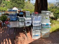 Full lot of 28 assorted plastic storage containers stacked on and beside a table outdoors.